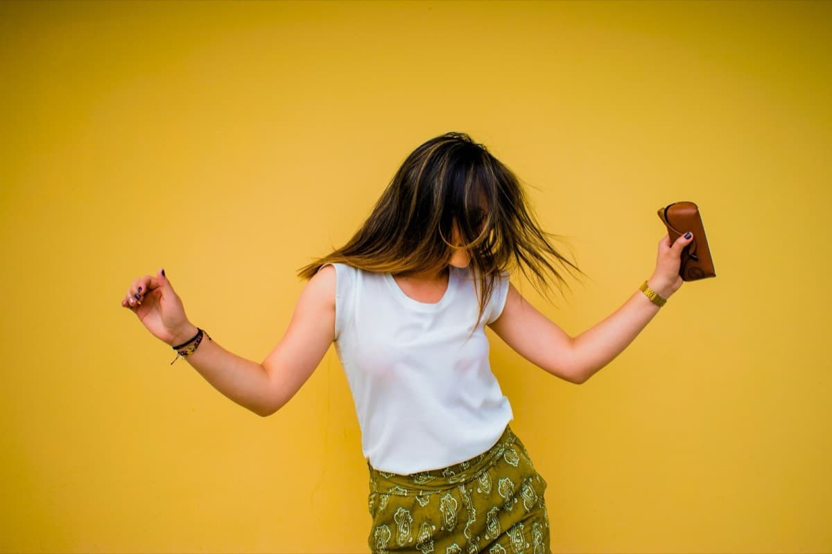 Someone dancing in front of a bright yellow wall, hair thrown across their face