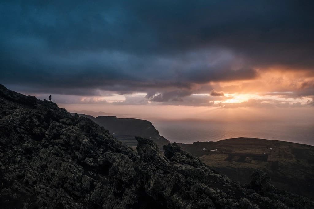 A walker standing on a dark volcanic ridge as the sun breaks under storm cloud over the sea