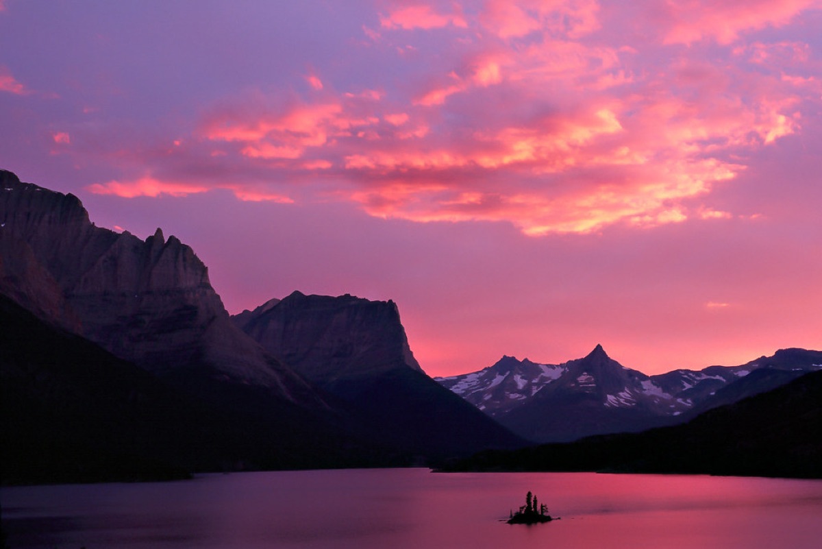 A pink and violet sunset over dark mountain silhouettes and a still lake
