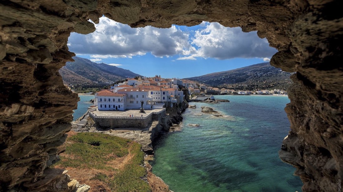 A white and terracotta coastal village on a headland, seen through a gap in a rock arch, turquoise water below