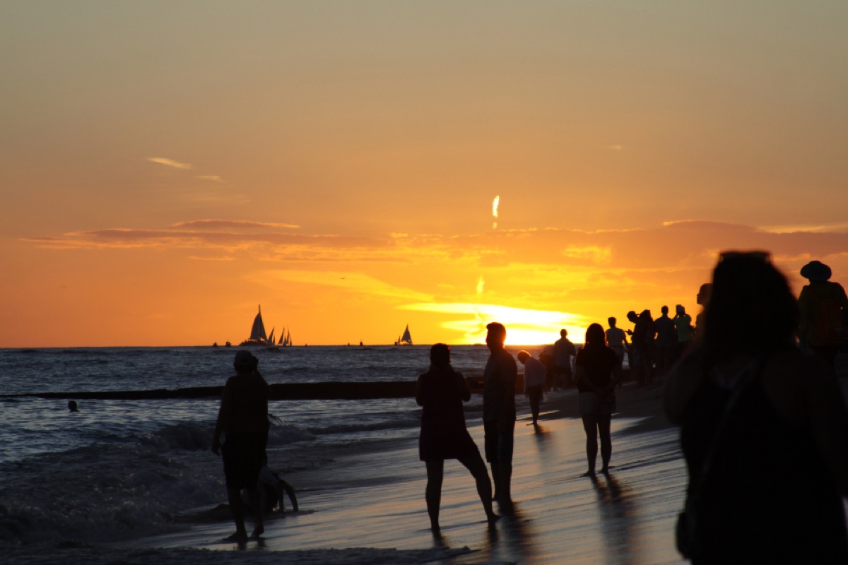 A crowd of silhouettes on a shoreline at dusk, with sailboats on the horizon