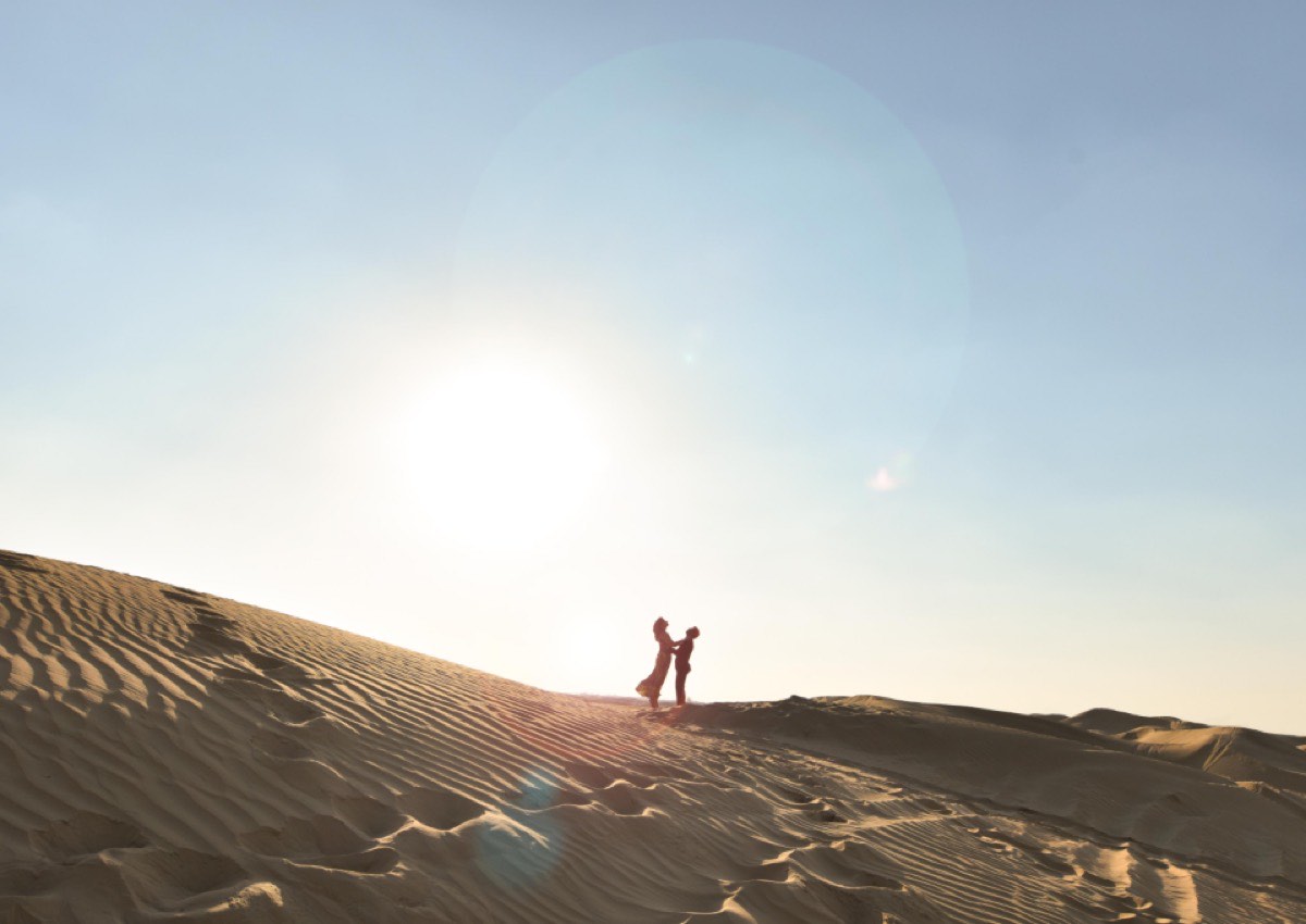 A couple silhouetted on a dune crest against a wide sunburst, ripples in the sand below