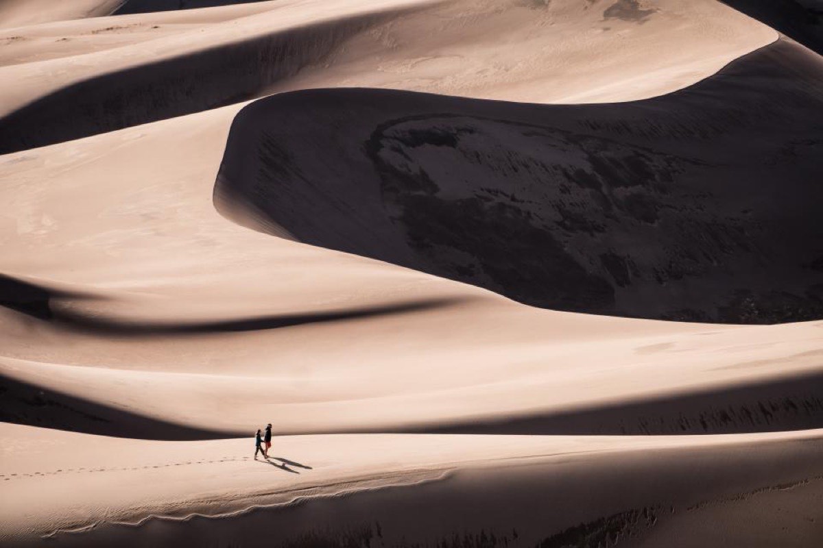 Two tiny figures crossing a sunlit sand ridge, long shadows behind them