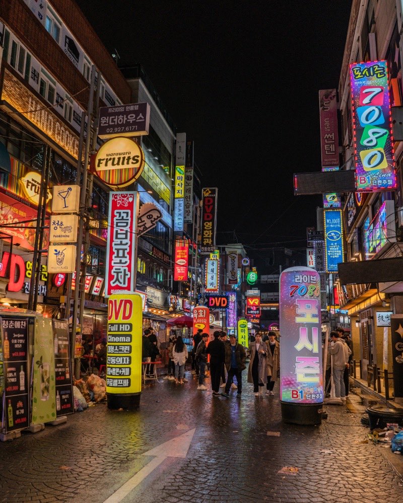 A night street stacked with pink, green and blue neon signs, people walking the wet centre