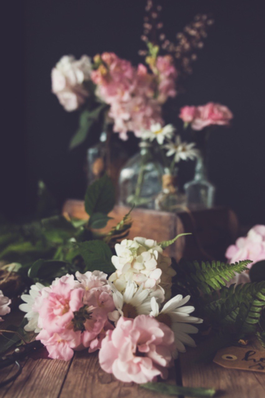A dark still life of pink and cream blooms arranged across glass bottles on a wooden crate