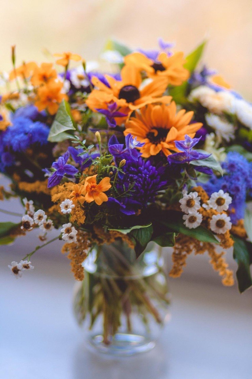 A dense posy of orange rudbeckia, blue cornflowers and white daisies in a clear glass vase