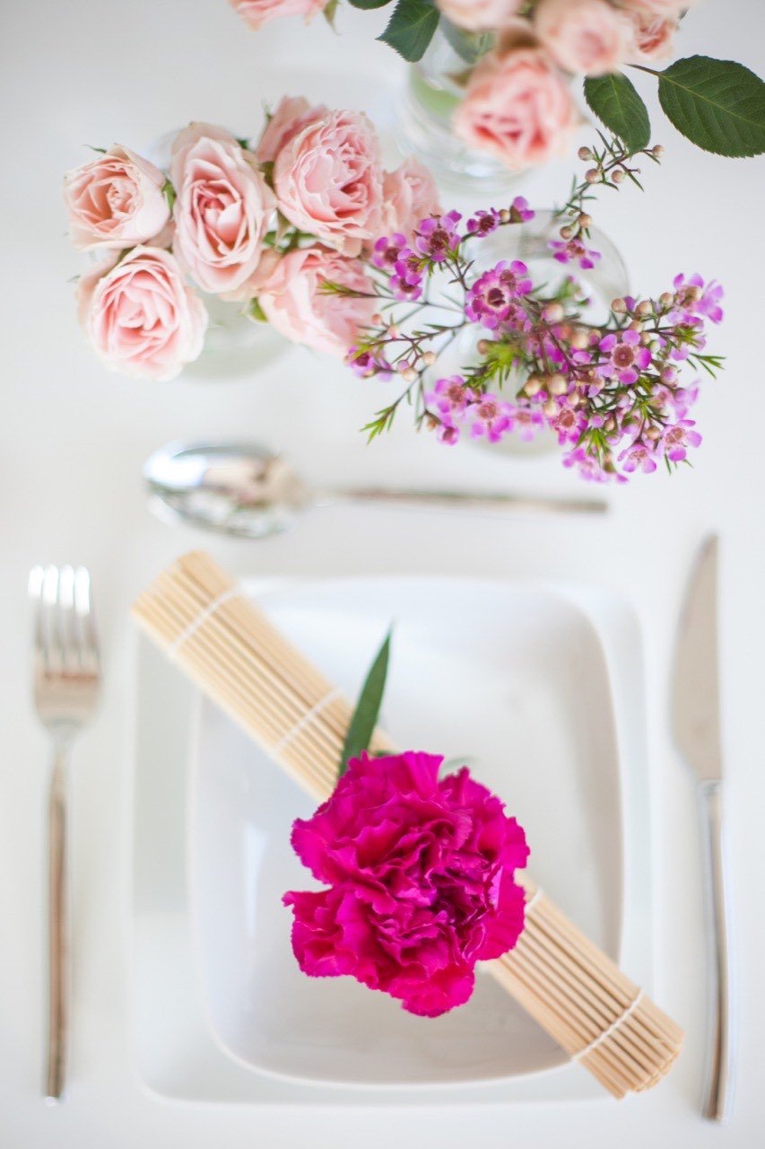 A white place setting with pale pink roses above it and a magenta carnation on a folded band