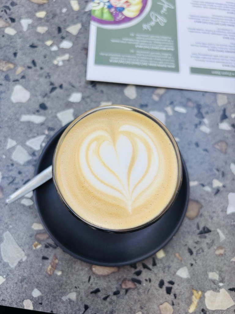 A flat white in a dark ceramic cup on a pale terrazzo table, photographed from above.