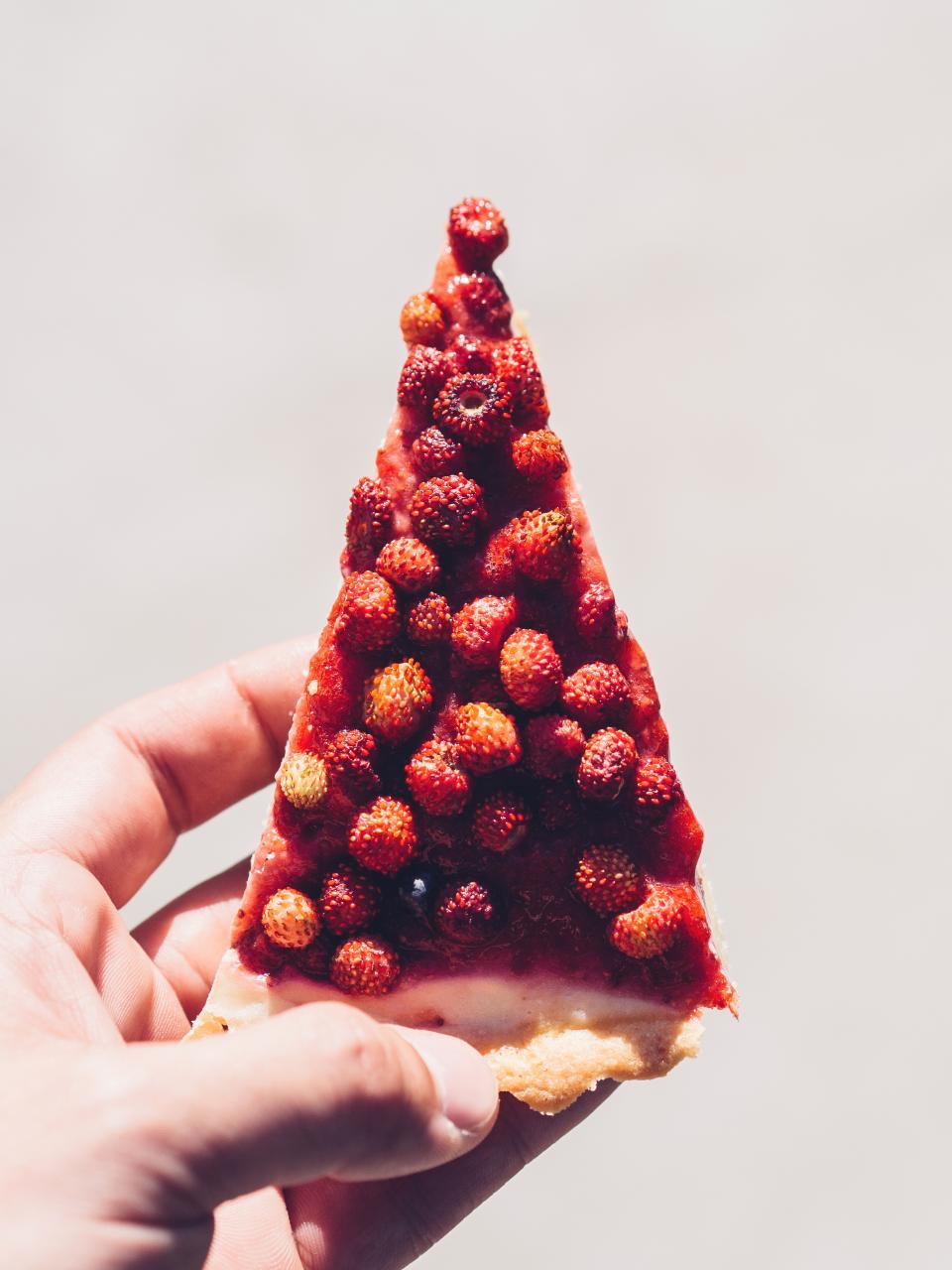 A hand holding up a wedge of tart packed edge to edge with wild strawberries, against a plain pale wall