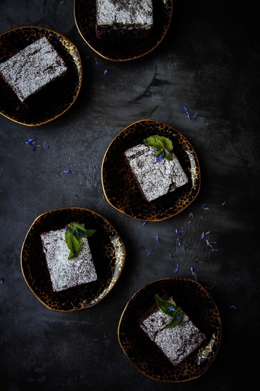 Five dusted brownie squares on small dark gold-rimmed plates scattered across a near-black surface with blue cornflower petals