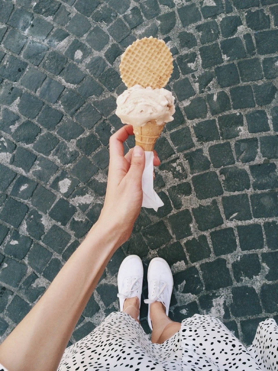 A first-person shot looking down at a hand holding an ice cream cone over grey cobblestones, the photographer’s own shoes at the bottom of the frame