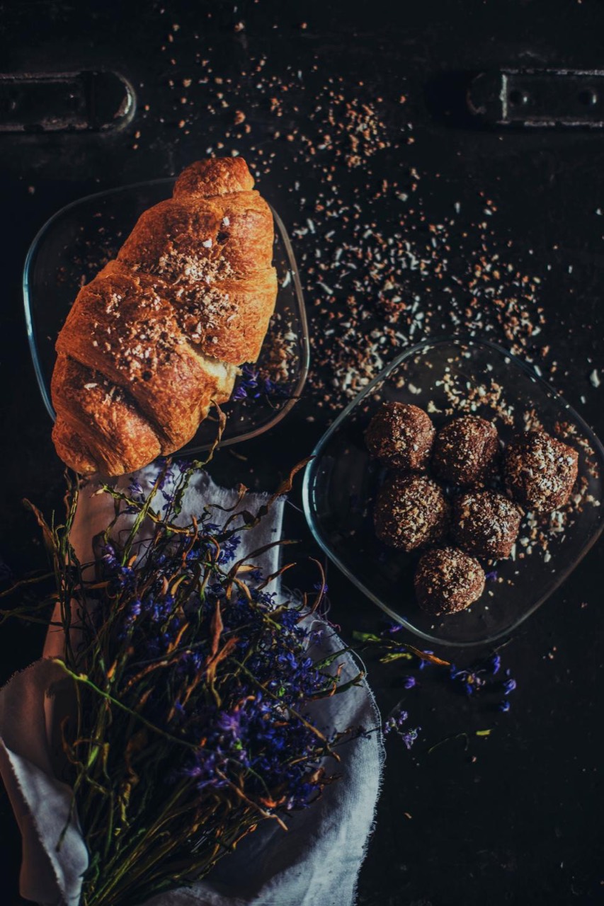 A croissant and a dish of cocoa truffles on a dark surface, dried flowers laid beneath and crumbs thrown across the frame