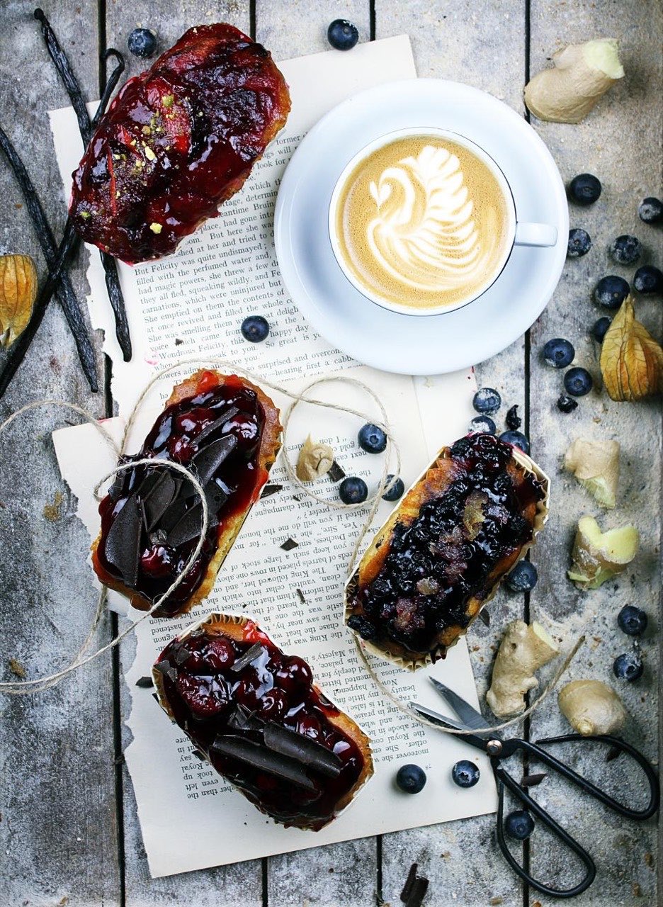 Four glazed éclairs laid on torn book pages with a cappuccino, vanilla pods, blueberries and a pair of scissors, shot from above