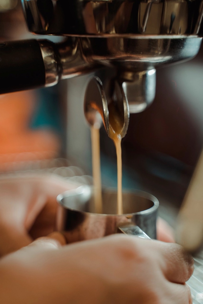 Espresso running from a naked portafilter into a steel pitcher, shot close and warm with hands at the edge of the frame