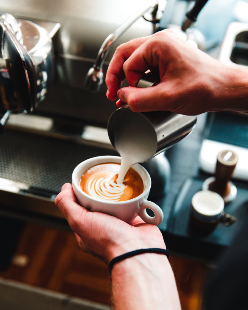 A cup held in one hand while the other pours a rosetta into it, the espresso machine group heads out of focus behind