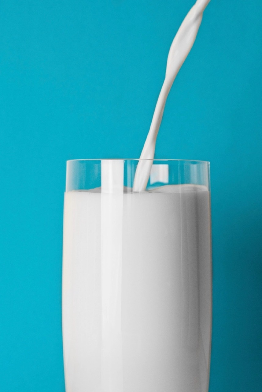 Milk pouring into a tall glass against a flat teal background, the stream frozen mid-fall