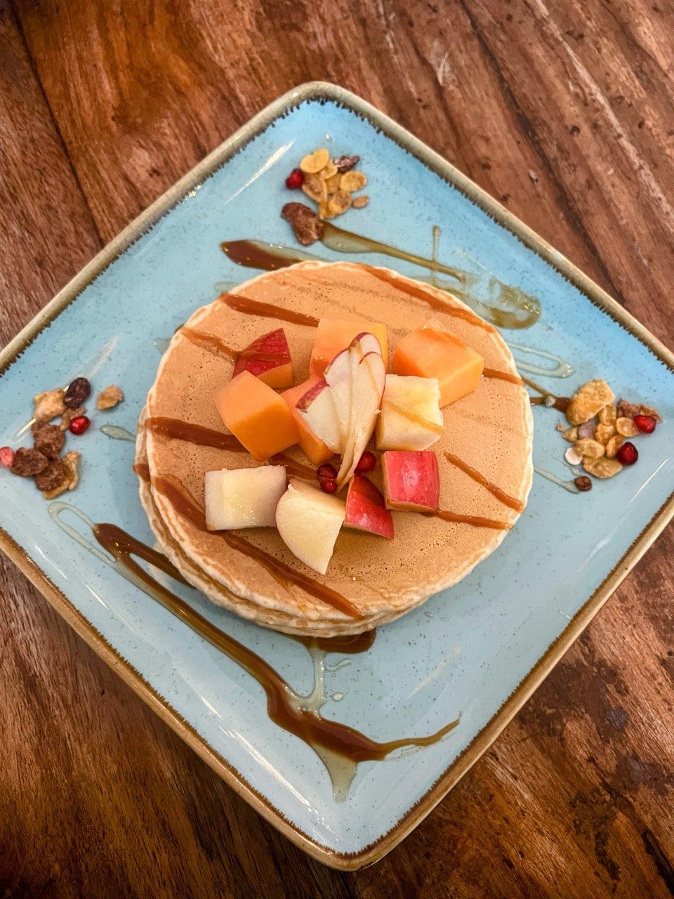 A short stack of pancakes on a square pale-blue plate topped with cut apple and melon and drizzled with caramel, shot from above on dark wood