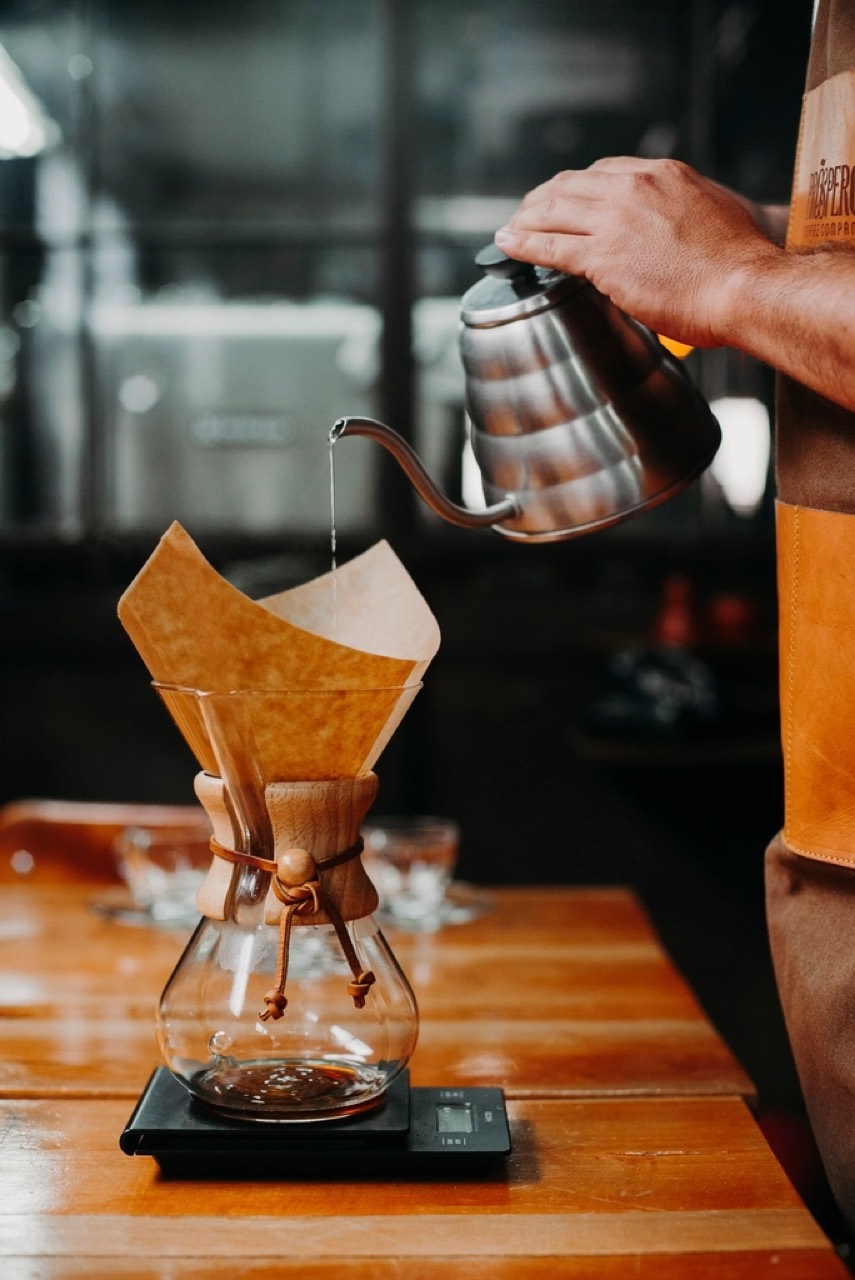 A gooseneck kettle pouring into a filter cone standing on a scale, the counter in warm wood and the room dark behind