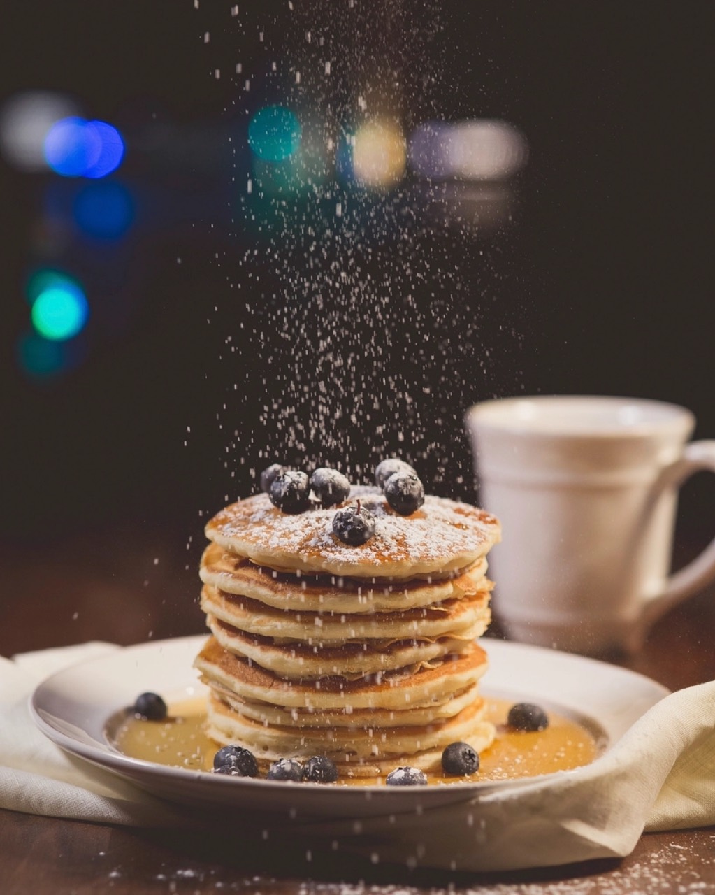 Icing sugar falling onto a tall stack of blueberry pancakes, coloured bokeh lights across the dark background