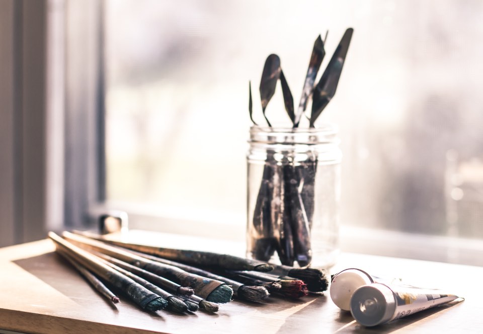 Palette knives standing in a glass jar on a windowsill, brushes laid out in front of it.