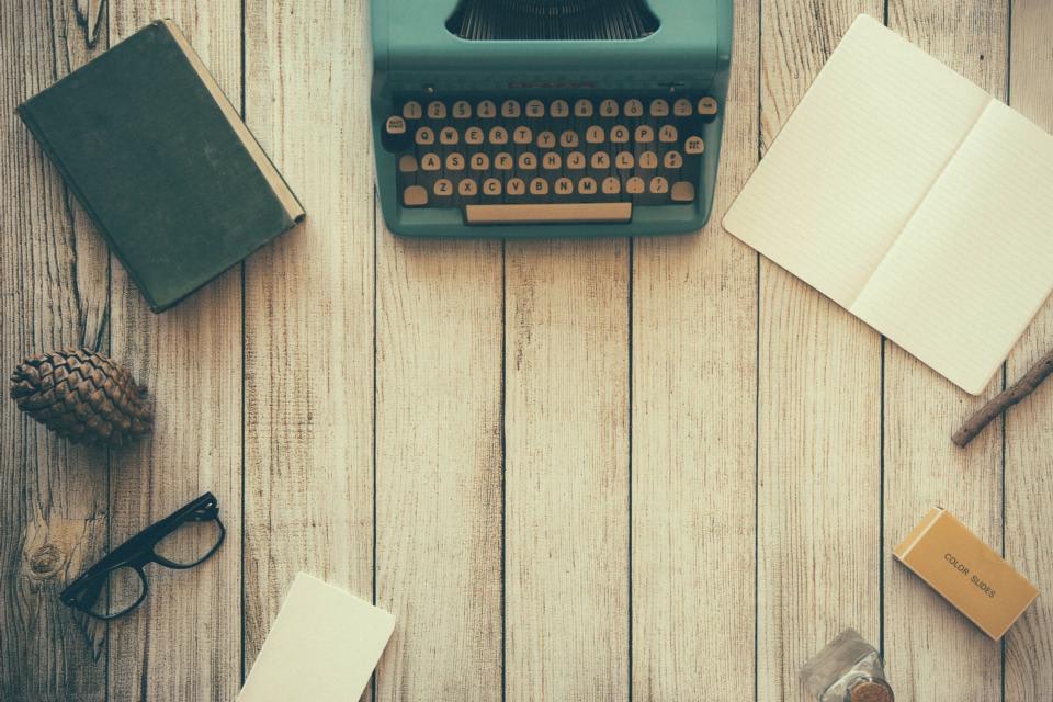 A mint green typewriter on pale boards, ringed by notebooks, reading glasses and a pine cone.