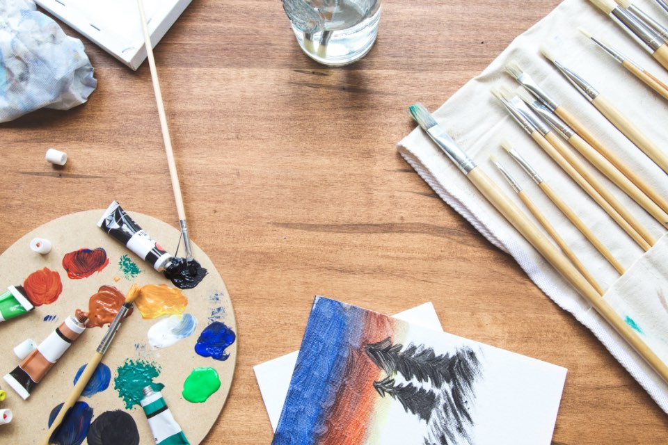 A paint palette loaded with colour, brushes and small prints spread across a wooden table.