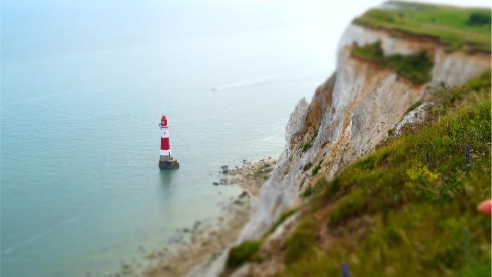 A red and white lighthouse standing in shallow green water below a grass-topped chalk cliff.