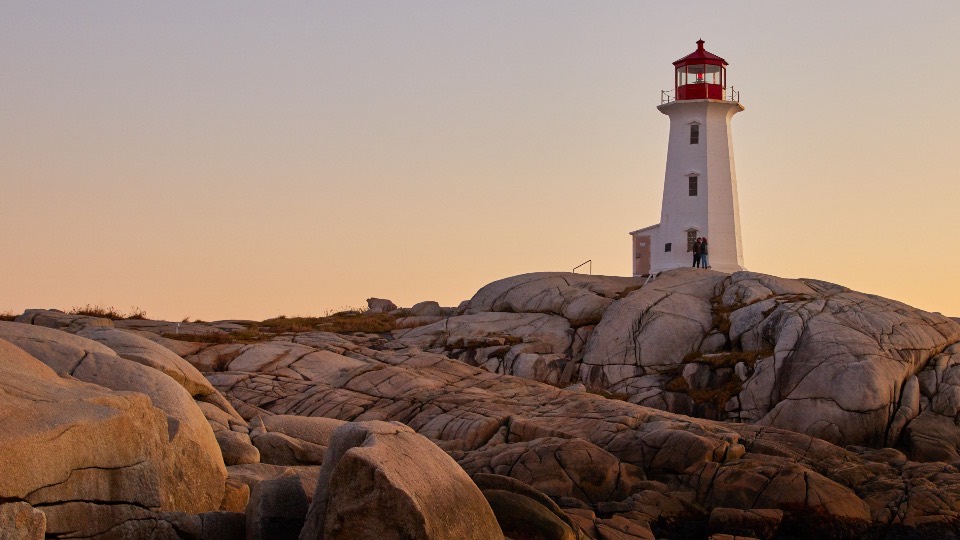 A white lighthouse with a red lantern room standing on smooth granite boulders at sunset.