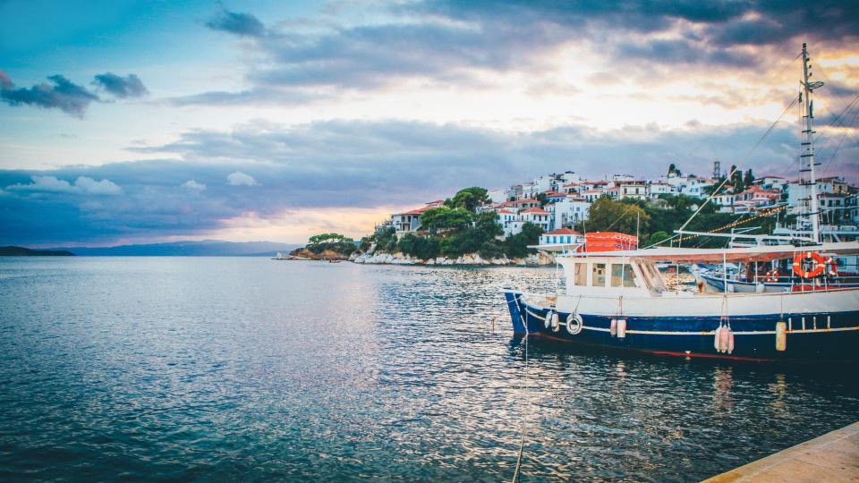 A white hillside village above a still bay, a blue and white fishing boat moored in the foreground.