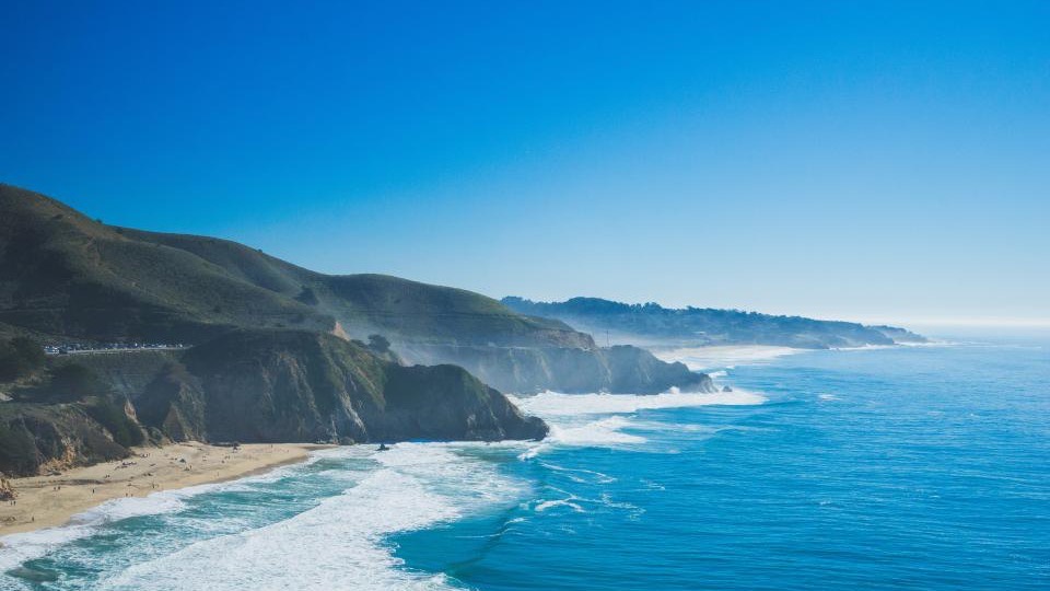 A long green headland dropping to a turquoise sea, surf lines running down the beach below.