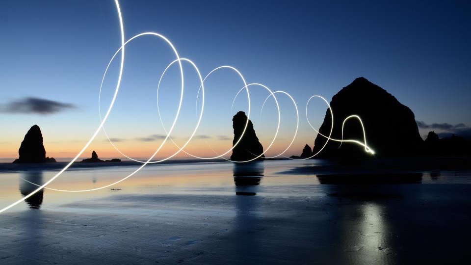 Sea stacks at dusk with a loop of light traced across the sky and reflected on the wet sand.