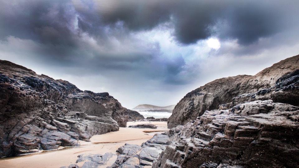 A rocky shore under a dark storm front, pale sand running between two shelves of layered rock.
