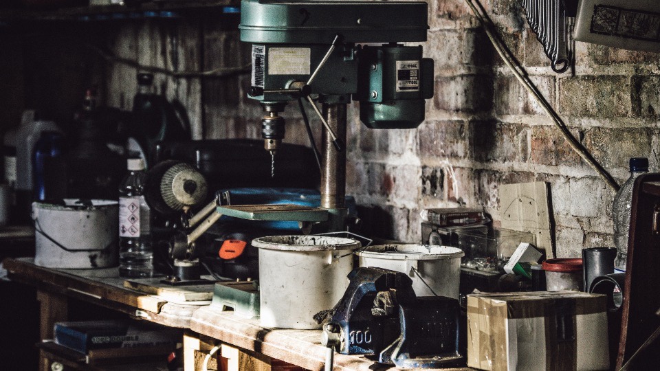 A pillar drill on a cluttered workbench against bare stone, tins and hand tools crowding the bench.