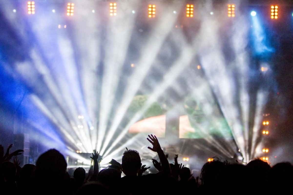 White and blue beams fanning out over an audience from a lighting rig, raised hands in silhouette along the bottom of the frame