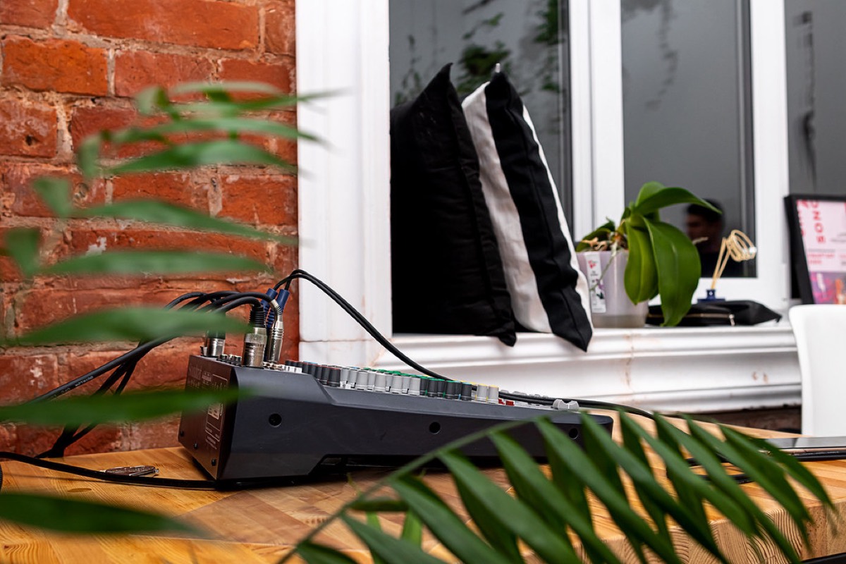 A small mixing desk patched with blue cables on a wooden table against a bare brick wall, a plant leaning into the frame