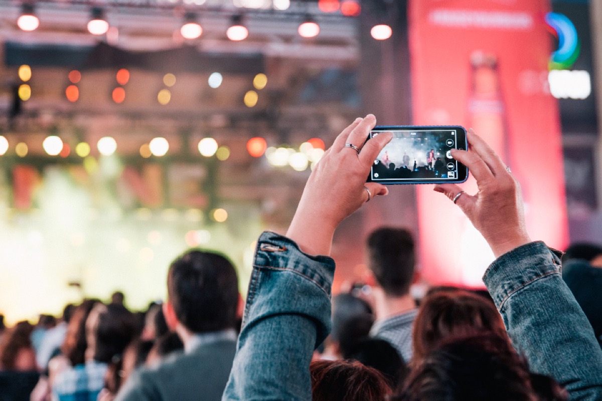 Two hands holding a phone up to film a lit stage, the screen showing the shot being taken, the crowd out of focus in front