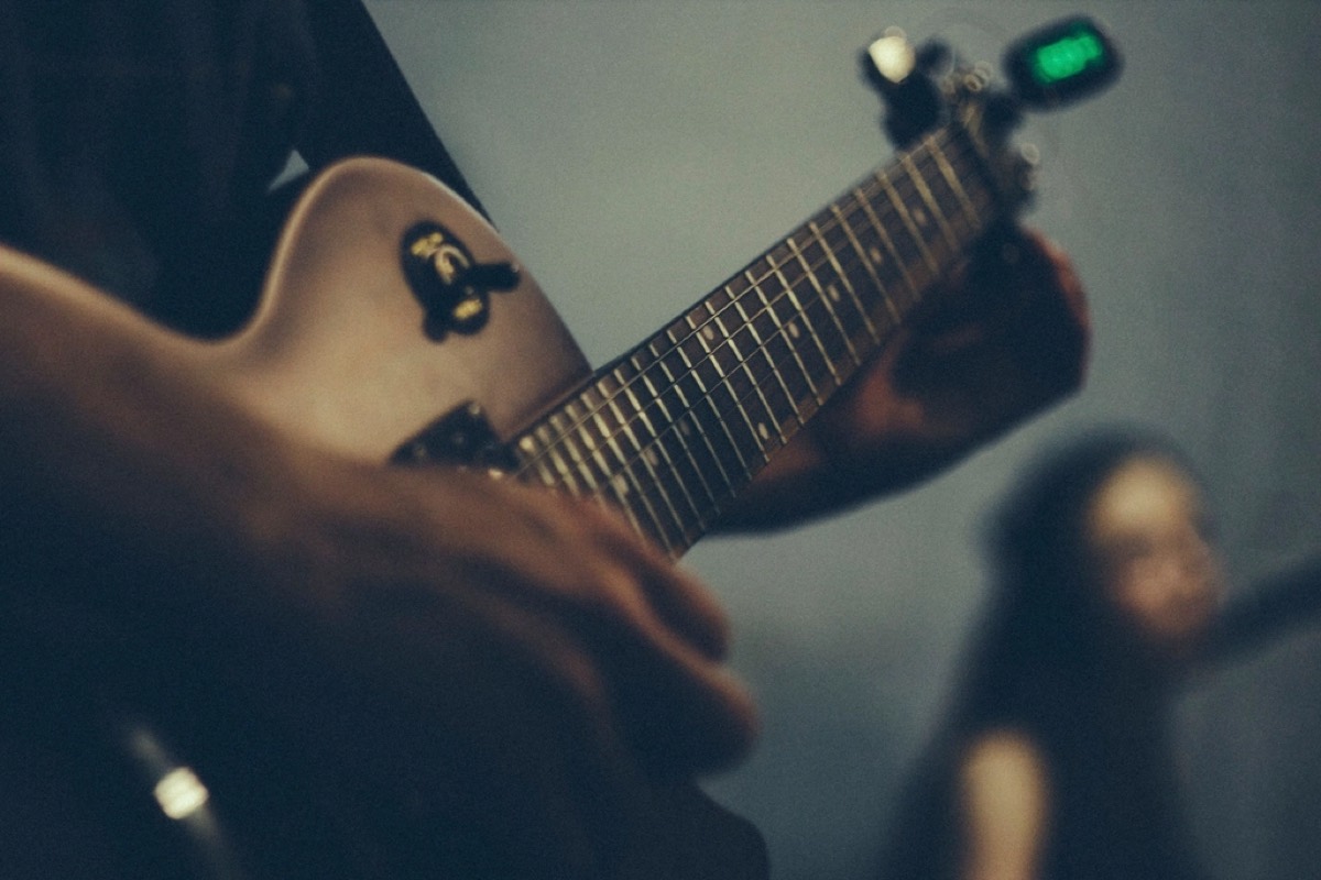 A close crop of a hand fretting an electric guitar, a clip-on tuner lit green on the headstock, the room out of focus behind