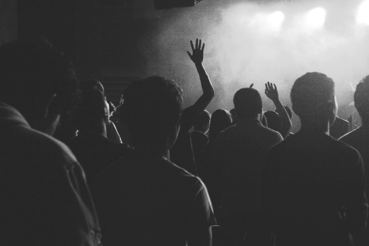 A crowd photographed from within, backs and raised hands in silhouette against a bright smoky stage, in black and white