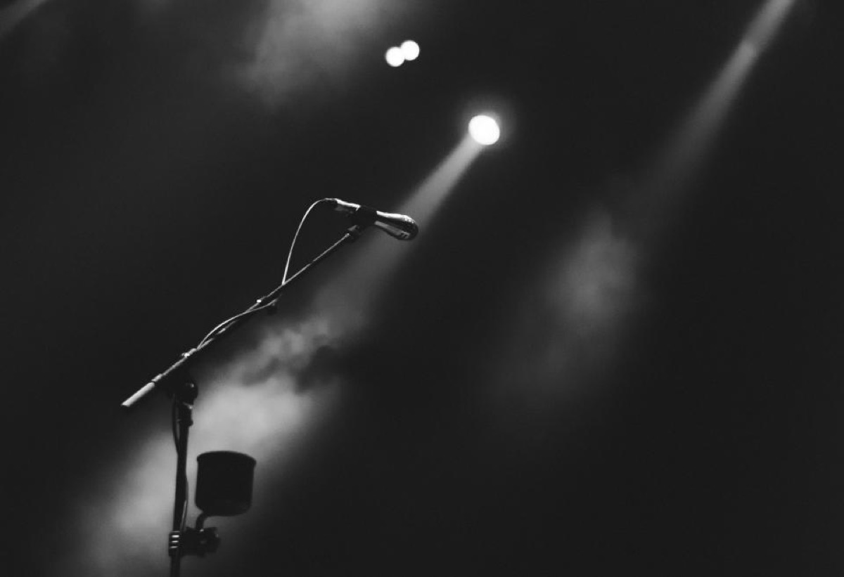 A vocal microphone on a boom stand caught in a single hard spotlight, smoke drifting through the beam, in black and white