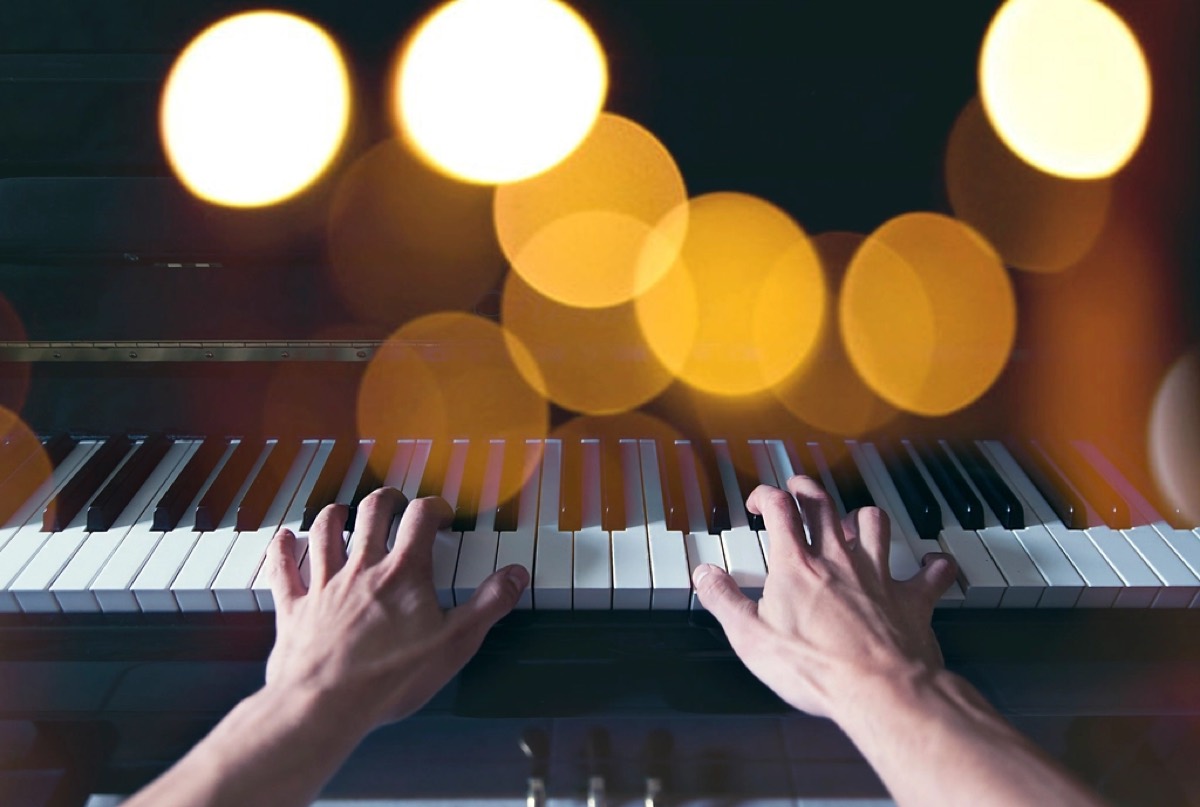 Two hands playing an upright piano, shot low across the keys, with large amber bokeh lights filling the top of the frame