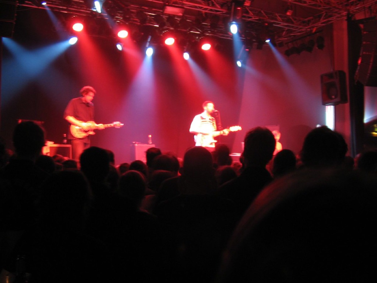 A small club stage under red and white par cans, two guitarists at mid distance, the crowd a dark silhouette across the lower half